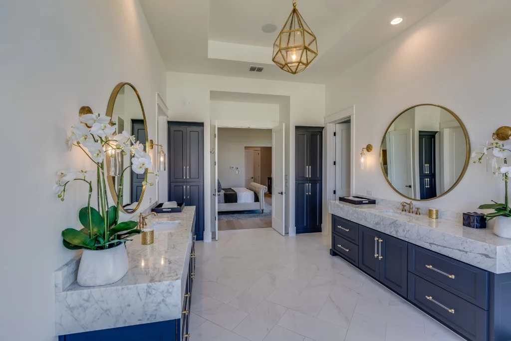 Elegant Double Vanity Bathroom with Marble Countertops Elegant bathroom featuring double vanities with marble countertops, round mirrors, dark blue cabinetry, gold fixtures, and a central chandelier, leading to a view of the adjoining bedroom.