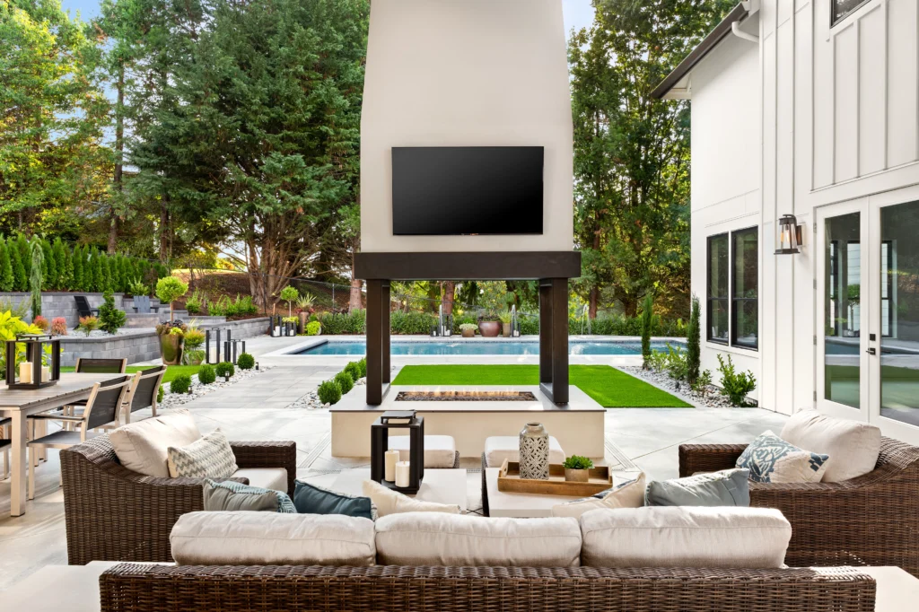 Luxurious Outdoor Living Space with Fireplace and Pool Beautifully designed outdoor living space featuring a cozy seating area with wicker furniture, a modern fireplace with a mounted TV, and a stunning pool surrounded by lush greenery.