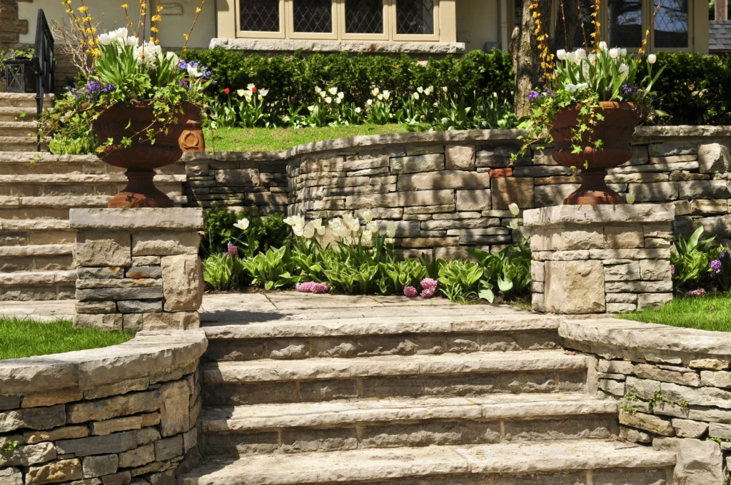 Elegant Stone Retaining Wall with Flower Beds Beautiful stone retaining wall with integrated steps, adorned with vibrant flower beds and large decorative planters, creating a charming and picturesque garden entrance.